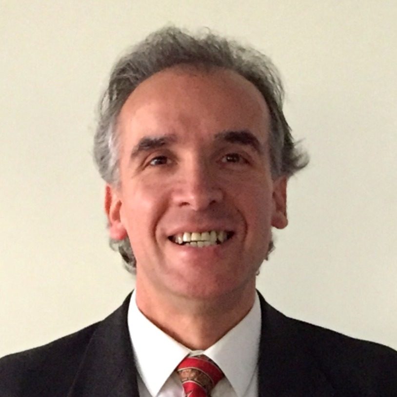 Smiling middle-aged man with gray hair in a dark suit and red tie, standing against a light background (professional portrait).