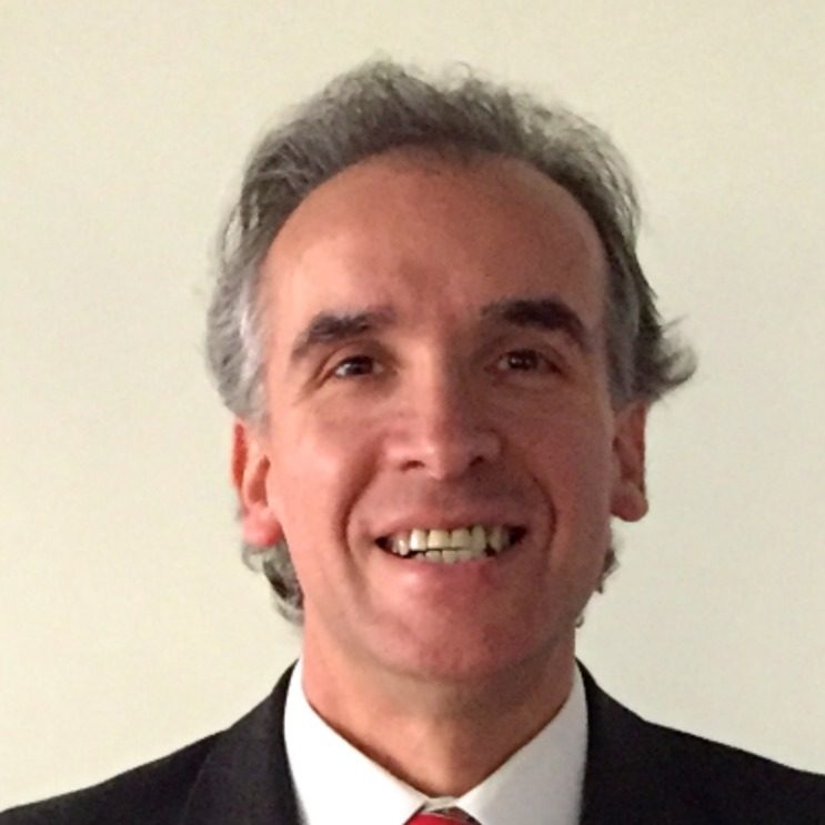 Smiling middle-aged man with gray hair in a dark suit and red tie, against a light background. There is a portrait-style head-and-shoulders shot with a friendly expression.